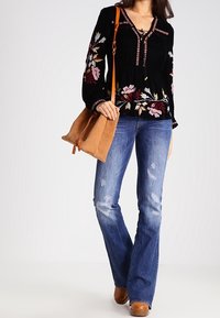 Blouse en velours noir avec broderies florales, associée à un jean flare bleu déchiré. Le modèle tient un sac à épaule en daim beige. Chaussures marron.