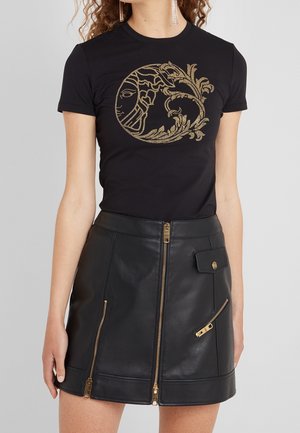Kobieta nosząca czarny t-shirt ze złotym motywem słońca i kwiatów, w połączeniu z czarną skórzaną spódnicą z złotymi suwakami i kieszenią na zatrzask.