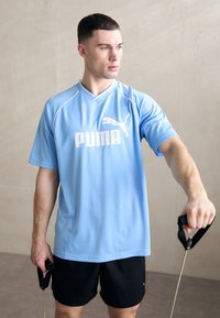 Lyst blå PUMA træningsshirt med hvidt logo, parret med sorte shorts. Den enkelte holder træningsbånd i en neutral setting.