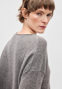 Szary dzianinowy sweter o luźnym kroju, z szerokim dekoltem i teksturowaną tkaniną. Sweter posiada delikatne prążkowane detale na rękawach.