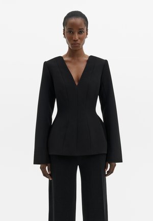 Donna in piedi che indossa un blazer nero aderente con scollo a V profondo e spalle strutturate, abbinato a pantaloni neri a gamba larga.