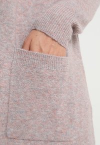 Pull à manches longues, de couleur rose pâle, avec une texture douce, doté d'un poignet côtelé et d'une poche à l'avant. Le tissu présente un motif légèrement moucheté.