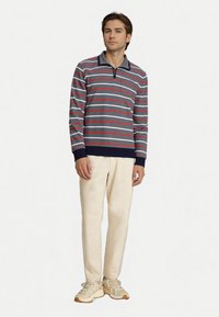 Langärmliges graues Polohemd mit roten und weißen Streifen, Reißverschlusskragen, linke Brusttasche, kombiniert mit hellbeigen Hosen und cremefarbenen Sneakers.