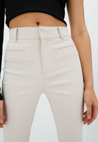 Pantalon beige ajusté avec une coupe cintrée, des détails discrets de poches et un devant plat. Le tissu est lisse, créant une silhouette épurée.