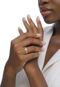 Bague en diamant en forme de cœur avec une bande délicate ornée de petits accents. Les mains sont légèrement manucurées avec des ongles naturels, sur un fond blanc.