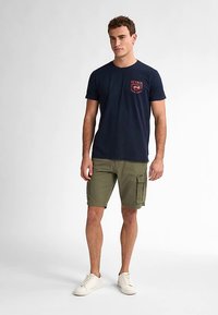 Tmavě modré tričko s červeným potiskem na kapse spojené s olivově zelenými kraťasy typu cargo. Stylizováno s bílými teniskami, stojící proti neutrálnímu pozadí.