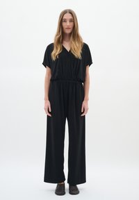 Svart jumpsuit med V-ringning, korta ärmar och elastisk midja. Har vida ben. Modellen bär mörka slip-on skor.