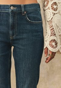 Los pantalones vaqueros de mezclilla azul oscuro presentan un diseño clásico de cinco bolsillos con herrajes de latón y un leve desteñido. Una manga de ganchillo es parcialmente visible.