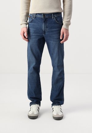 Person wearing blue straight-leg jeans, waffle-knit beige sweater, and white sneakers with black stripes, standing on white floor.