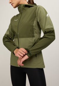 Olijfgroene jas met een ritssluiting aan de voorkant, verstelbare manchetten, zijzakken en logo-detail op de mouw. Gladde, lichte stof.
