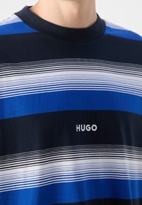 Gros plan d'une personne portant un t-shirt rayé bleu marine, bleu et blanc avec le logo « HUGO » brodé sur la poitrine.