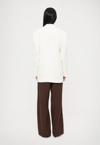 Biely nadrozmerný blejzer s klapákmi s výrezmi a gombíkovými manžetami, kombinovaný s hnedými širokými nohavicami. Hladká textúra, minimalistický dizajn.