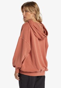 Felpa oversize in terracotta con cappuccio regolabile, polsini a costine e vestibilità rilassata. Texture morbida con dettagli di cucitura sulla parte posteriore.