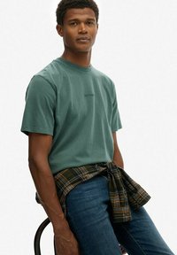 Zielona bawełniana koszulka z krótkim rękawem, z dekoltem w serek i wyszywanym logo. Koszula w kratę związana w talii, niebieskie dżinsy, siedząca.