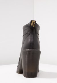 Černá kožená kotníková bota s vysokým, vroubkovaným podpatkem. Má polstrovaný límec a malou poutko se žlutým textem vzadu.