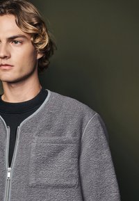 Jachetă de fleece gri cu fermoar frontal, cu manșete ribbate și un buzunar pe piept. Are un gât rotund și detalii cu cusături contrastante.