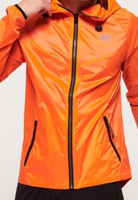 Personne portant une veste à capuche orange vif avec des détails de fermeture éclair et de poches noires, ajustant le col d'une main.