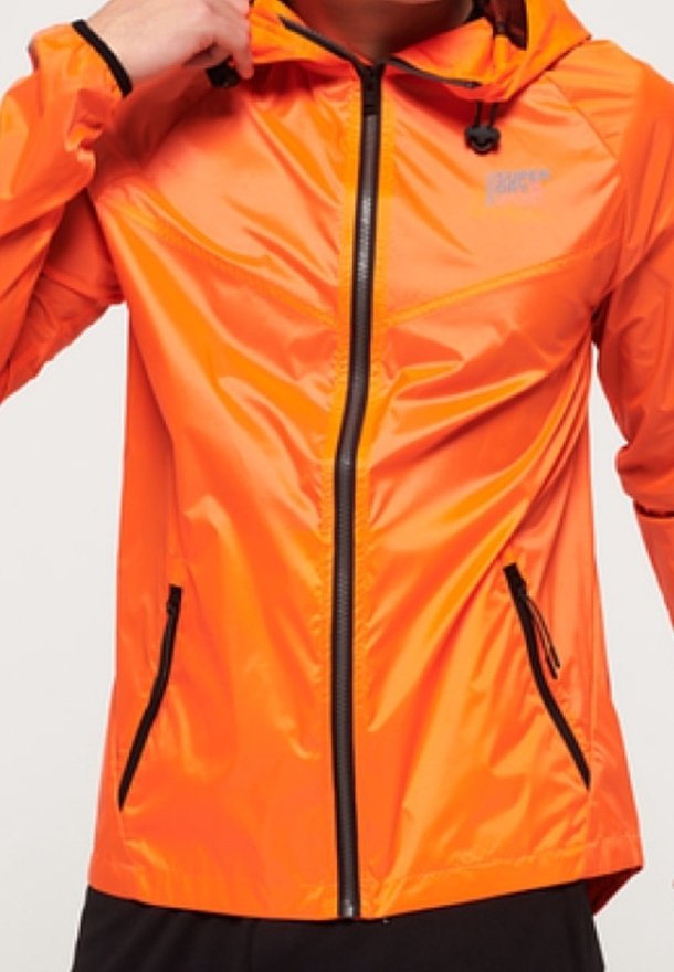 Personne portant une veste à capuche orange vif avec des détails de fermeture éclair et de poches noires, ajustant le col d'une main.