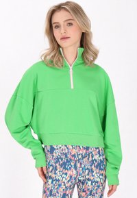 Zöld pulóver, puha anyagból készült, félcipzárral és ezüst karikával rendelkezik, valamint cropped dizájnnal bír. Tarka mintás leggings-szel párosítva.