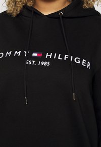 Black hoodie with "Tommy Hilfiger Est. 1985" embroidered in white and red, white, and blue logo on chest, worn by person with curly blonde hair.