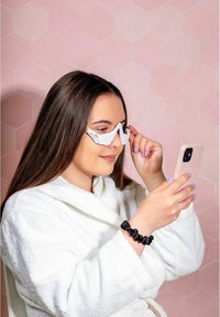 Witte, hoekige zonnebril met doorzichtige glazen, gedragen door een persoon in een witte robe. Houdt een lichtkleurige smartphone vast tegen een roze hexagonale achtergrond.