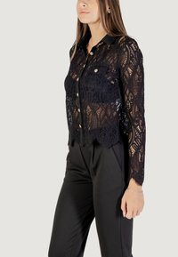 Camicia nera in pizzo con maniche lunghe, chiusura frontale con bottoni e colletto. Il motivo intricati e l'orlo scallop aggiungono dettaglio. Completata da pantaloni scuri.