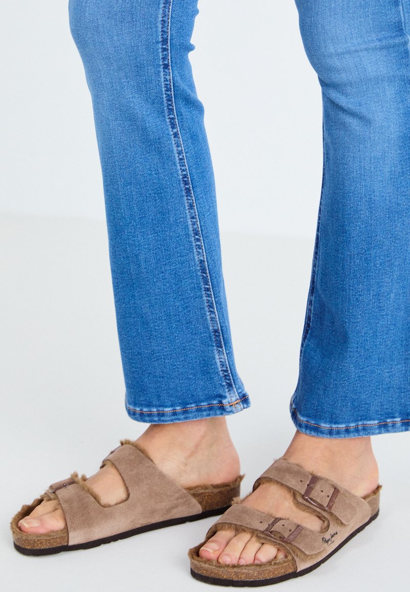 Braune Wildleder-Slidesandalen mit zwei verstellbaren Riemen, pelzähnlichen Akzenten und einer Korksohle, kombiniert mit hellblauen Schlagjeans.