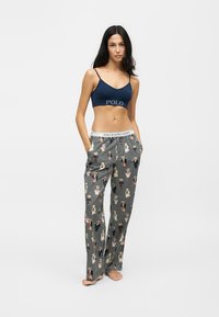 Femme portant un débardeur court bleu marine et un pantalon de pyjama rayé avec des motifs d'ours, debout pieds nus, les mains dans les poches.