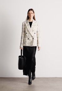 Διπλής όψεως υφασμάτινο σακάκι σε κρέμ απόχρωση με πολύχρωμες κλωστές, συνδυασμένο με μαύρο φόρεμα και ψηλές μαύρες μπότες, κρατώντας μια μαύρη τσάντα tote.