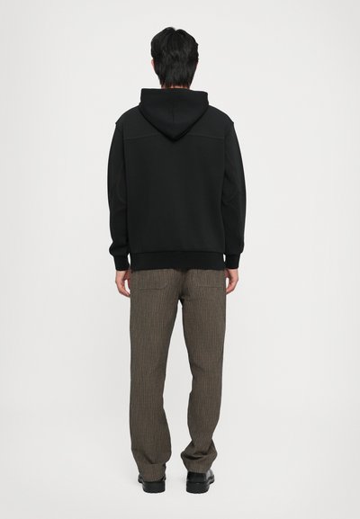 Felpa nera con cappuccio e cuciture a contrasto, abbinata a pantaloni a righe sottili marroni e scarpe nere. Modello mostrato di spalle.
