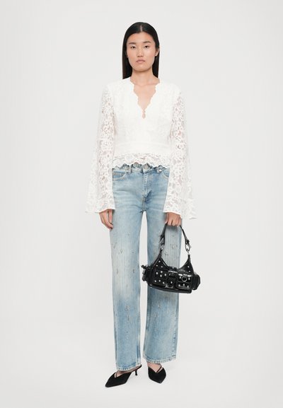 Witte kanten blouse met een golvende onderrand en een keyhole-halslijn, gecombineerd met lichtblauwe versleten jeans versierd met metalen accenten. Zwarte handtas.