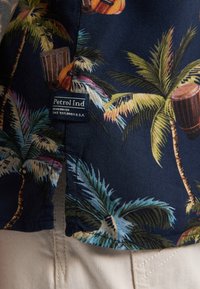 Camicia a maniche corte in tessuto blu scuro con motivi di palme tropicali e strumenti musicali; presenta un'etichetta "Petrol Ind" sull'orlo.