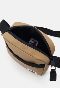 Bolso cruzado beige con cierre con cremallera, correa negra y bolsillo exterior. El interior cuenta con una etiqueta de logo y una textura suave.