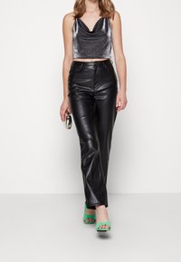 Pantaloni altissimi in pelle nera abbinati a un top corto a collo alto in metallo argentato. La modella indossa sandali verde con tacco e tiene in mano una pochette argentata.