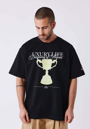 Man met kort haar en baard die een zwarte oversized T-shirt draagt met een trofee en tekst "Luxury Life Dreaming of Monaco."