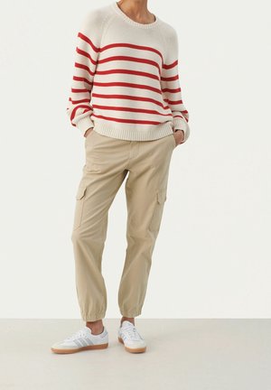 Personne portant un pull crème à rayures rouges, un pantalon cargo beige avec des poignets élastiques et des baskets blanches à semelles en gomme, debout les mains dans les poches.