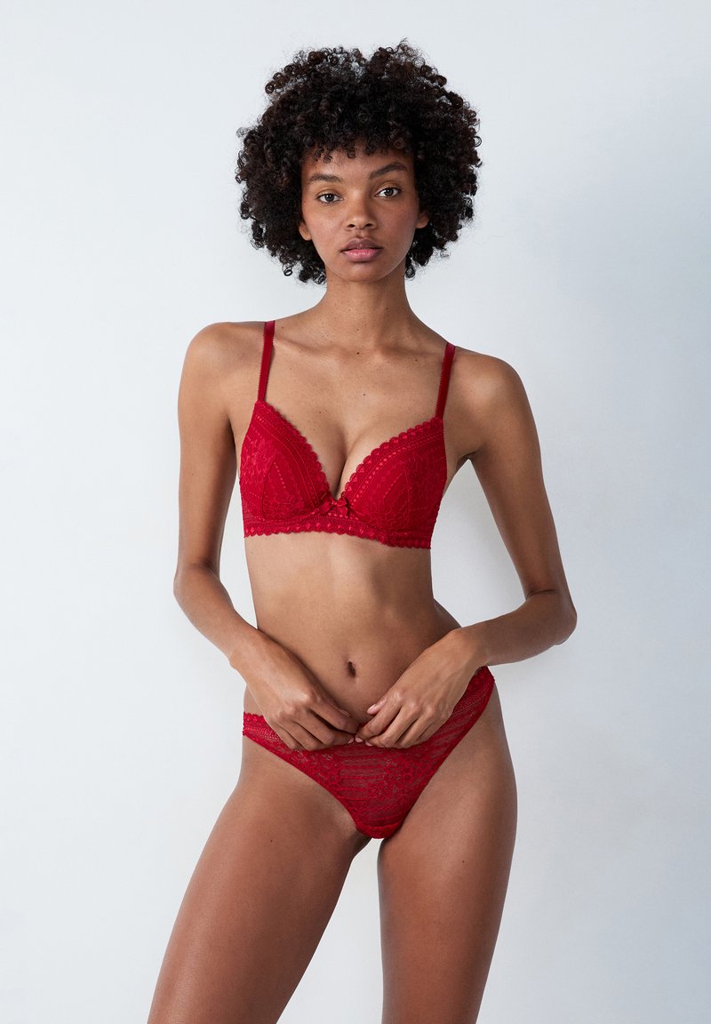 Červený krajkový bralet a tanga set s vlnkovými okraji, trojúhelníkovými košíčky a dekorativní mašlí ve středu. Hladká textura s květinovými vzory.