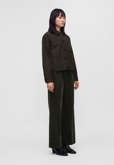 Femme debout portant une veste sombre à boutons, un pantalon ample et sombre à jambes larges, et des bottines noires, sur un fond clair uni.