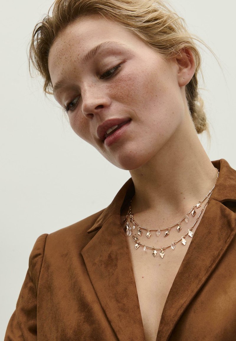 Collana a tre strati con catene in metallo dorato, caratterizzata da ciondoli a forma di foglia e accenti di gemme trasparenti, indossata sopra un blazer in suede marrone.