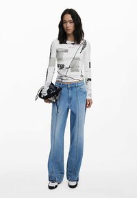 Wit shirt met lange mouwen met zwarte tekst en grafische prints, gecombineerd met losse blauwe spijkerbroeken en zwart-witte sneakers.