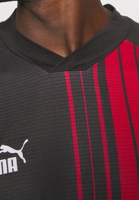 Jersey deportivo negro con franjas verticales rojas, tejido texturizado y diseño de cuello en V. Presenta un logo blanco de Puma en la parte inferior izquierda.
