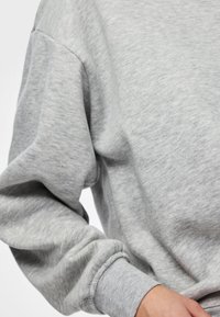 Artimos planas asmens, dėvinčio šviesiai pilką ilgomis rankovėmis džemperį su gofruotais rankogaliais, baltame fone.