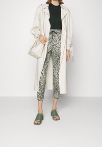 Lang, lichtbeige trenchcoat over een donkergroene top. Bedrukte groen en zwarte leoprint broek, gecombineerd met groene slidesandalen.