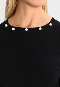 Svart ribbad topp med en vid halsringning och jämnt fördelade pärldetaljer runt kragen. Slät textur med en åtsittande design.