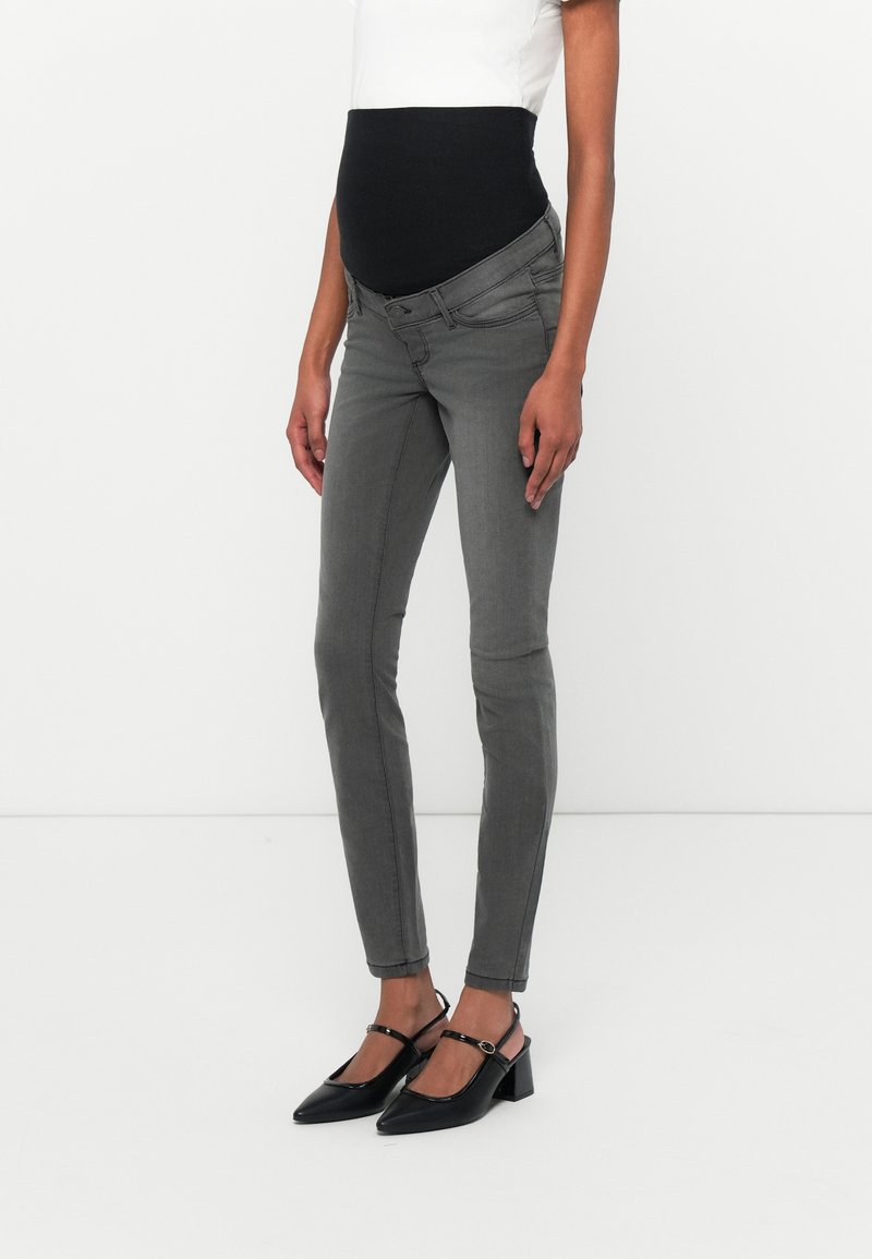 Maternity skinny jeans in grey, featuring a black elastic over-bump panel, five-pocket design, and slim-fit silhouette. Paired with black heels.