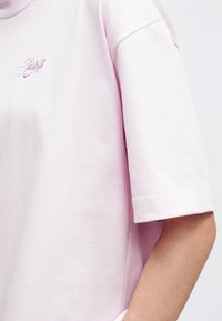 Άτομο που φοράει ένα ανοιχτό ροζ κοντομάνικο πουκάμισο με ραμμέν α γράμματα στο στήθος, στέκοντας μπροστά σε έναν απλό ανοιχτό φόντο.