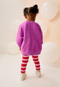 Paarse oversized sweatshirt, gestreepte rode en witte legging, en witte schoenen met textuurdetails, tegen een lichte achtergrond met ballonnen.