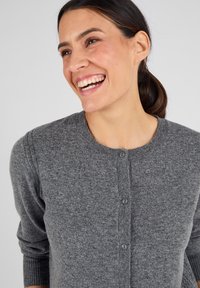 Cardigan gris en tissu texturé doux, doté d'un col arrondi et d'une fermeture à boutons avec des coutures visibles et des poignets côtelés.