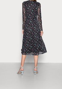 Schwarzes Blumenkleid mit langen, durchsichtigen Ärmeln, das eine Mischung aus blauen und rosa Blumen zeigt. Fließt bis unter das Knie und wird mit hellblauen, trägerlosen Absätzen kombiniert.