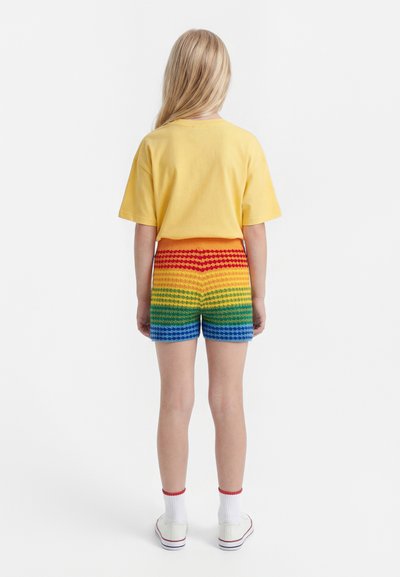 Enfant aux cheveux blonds longs portant un t-shirt jaune à manches courtes, un short rayé arc-en-ciel, des chaussettes blanches avec une bordure rouge et des baskets blanches, debout de dos.